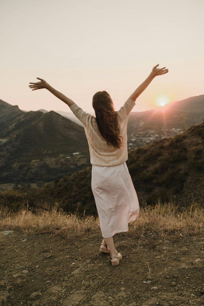 Une femme de dos, les bras levés, contemple un coucher de soleil dans un paysage de montagne.