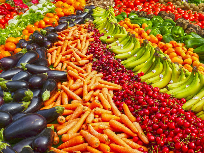 Étal de marché coloré avec fruits et légumes frais (carottes, bananes, cerises, tomates, aubergines, poivrons) illustrant une alimentation brute et variée.