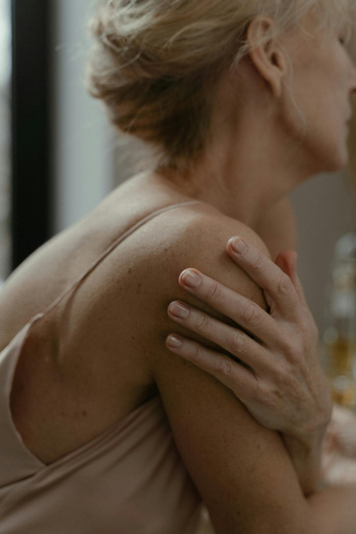 Gros plan élégant sur l'épaule et la main d'une femme d'âge mûr aux cheveux blonds attachés, portant un caraco en soie beige. Sa main repose doucement sur son épaule, suggérant le confort et l'autosoing après l'apaisement des douleurs articulaires ou de…