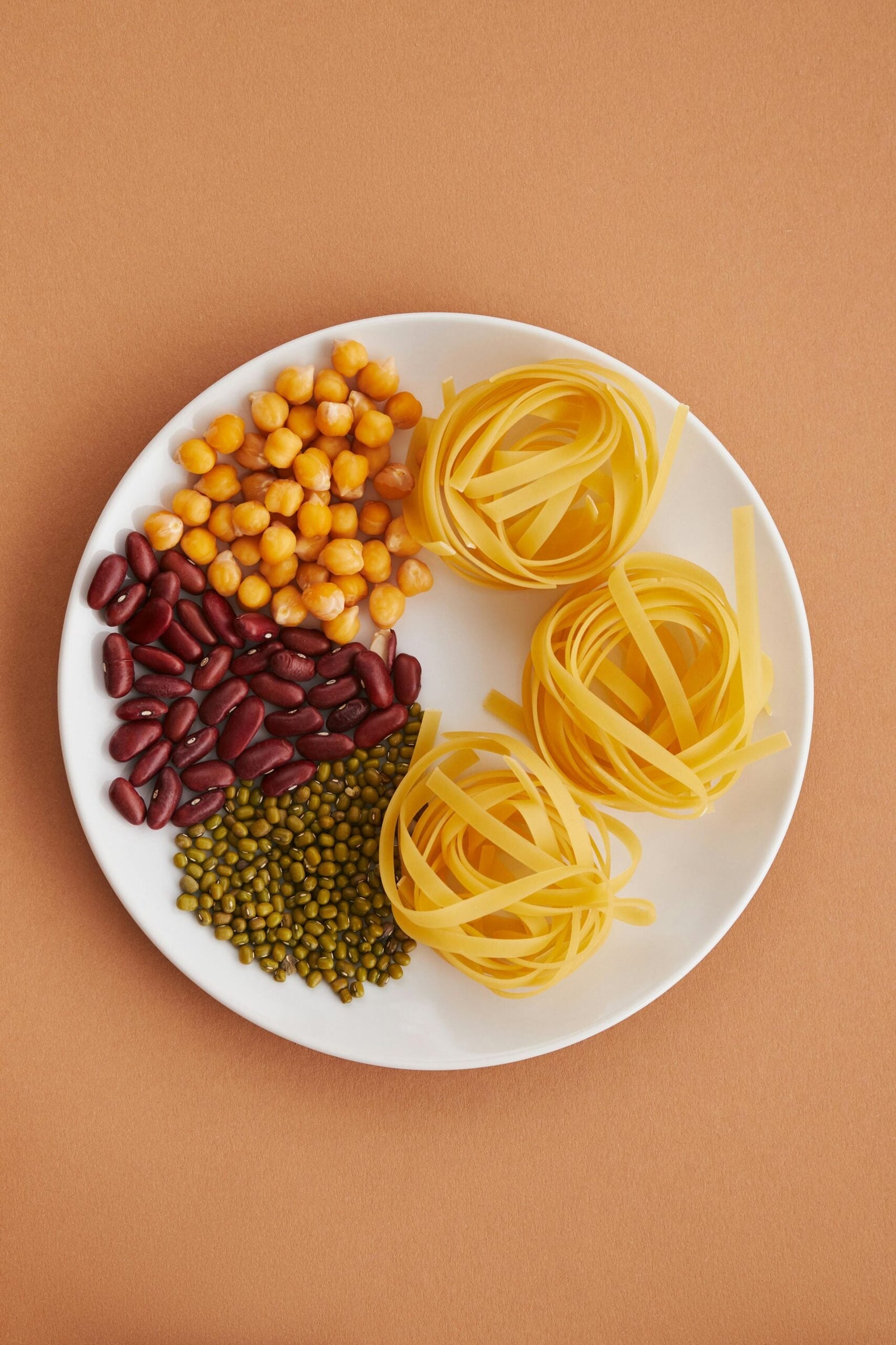 Assiette blanche vue du dessus avec des pois chiches, des haricots rouges, des haricots mungo et des nids de pâtes sur un fond beige.